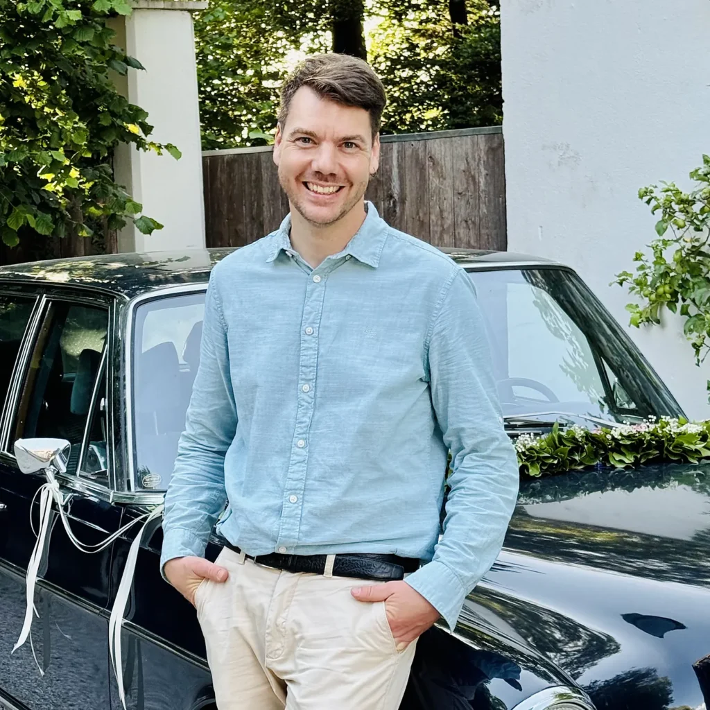 Chris, DJ aus München, lehnt lächelnd an einem geschmückten Oldtimer-Hochzeitsauto