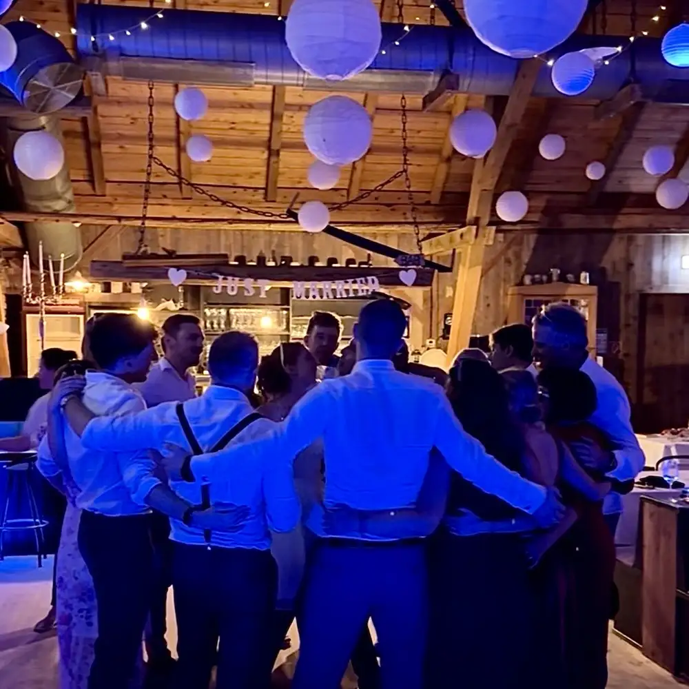 DJ New Inside: Hochzeitsgäste stehen Arm in Arm im Kreis auf der Tanzfläche der Alten Tenne in Moosinning, darüber Lampions und Just-married-Girlande