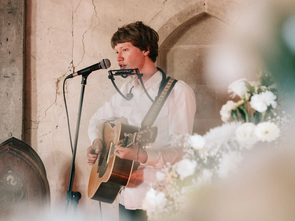 Musiker singt mit Akustikgitarre und Mundharmonika bei einer Trauung, im Vordergrund weiße Blumen